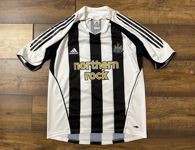 K&ouml;nnte beinhalten: Ein schwarz-wei&szlig; gestreiftes Fu&szlig;balltrikot mit dem Schriftzug "northern rock" in Gold. Das Trikot hat das Adidas-Logo und das Wappen von Newcastle United. Die &Auml;rmel sind wei&szlig; mit schwarzen Streifen.