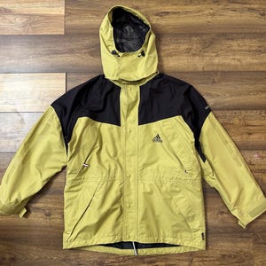 Puede incluir: Chaqueta con capucha Adidas amarilla y negra. La chaqueta tiene la parte superior del pecho y la capucha negras, con el cuerpo y las mangas amarillas. El logotipo de Adidas está en el pecho izquierdo. Tiene cremallera y bolsillos.