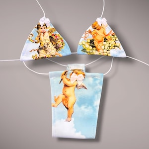 May include: A string of decorative fabric triangles and rectangles, each featuring a cherub design. The cherubs are depicted in a classical style, with wings and soft, warm skin tones, set against a light blue sky background. Each piece is adorned with a white bow.