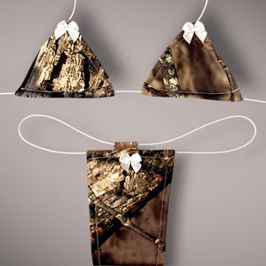 May include: Three camouflage fabric ornaments, two triangular and one rectangular, are suspended by white strings. Each ornament is adorned with a small white bow. The ornaments have a brown and green pattern.