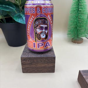 May include: A purple and orange can of Tennessee Brew Works "Hippies & Cowboys" India Pale Ale. The can features a cartoon of a man with a bandana and sunglasses. The text "IPA" is printed in large letters on the can.