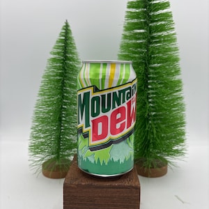 May include: A Mountain Dew soda can with a green, white, and yellow striped design, and the Mountain Dew logo in black and red. The can is on a small, dark brown wooden block, with two green bottle brush trees in the background.