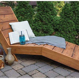 May include: A wooden chaise lounge with a gray and white blanket, a book, and a glass jar with a straw on top. The chaise lounge is on a patio with a brick walkway.