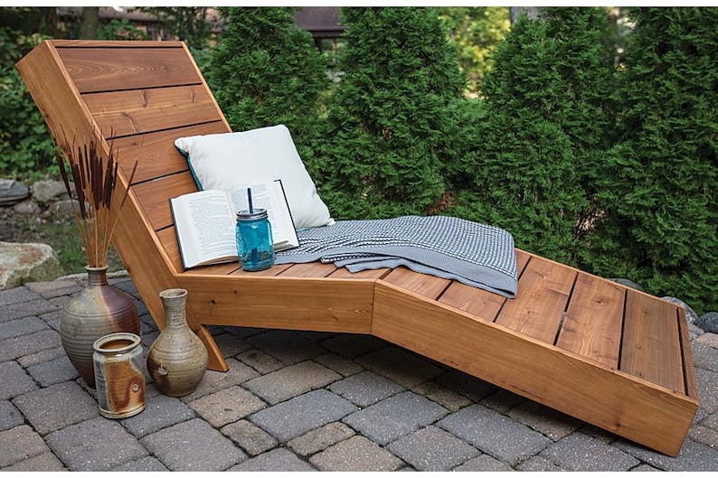 May include: A wooden chaise lounge with a gray and white blanket, a book, and a glass jar with a straw on top. The chaise lounge is on a patio with a brick walkway.