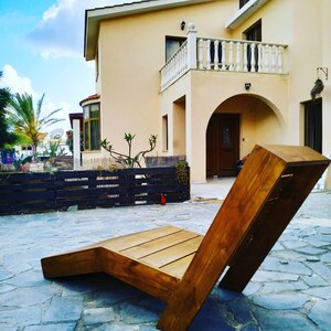 May include: A wooden lounge chair with a natural brown finish. The chair is made of thick planks of wood and has a simple, modern design. The chair is sitting on a patio in front of a house.