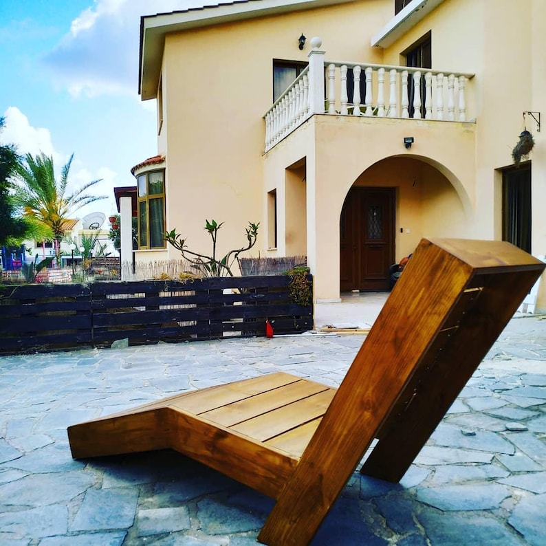 May include: A wooden lounge chair with a natural brown finish. The chair is made of thick planks of wood and has a simple, modern design. The chair is sitting on a patio in front of a house.
