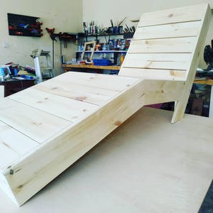 May include: A wooden lounge chair with a slatted back and a wide, angled seat. The chair is made of unfinished wood and is ready to be stained or painted.