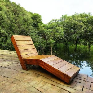 May include: A wooden lounge chair with a natural brown finish. The chair is made of slats and has a curved design. It is positioned on a brick patio overlooking a body of water.