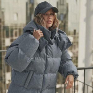 Puede incluir: Una chaqueta acolchada gris con gorra a juego. La chaqueta presenta un diseño con capucha, cierres de botón y bolsillos. El modelo lleva vaqueros azules. La chaqueta parece estar hecha de un material suave y texturizado.