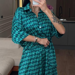Puede incluir: Un conjunto de dos piezas compuesto por una camisa de manga corta con botones y pantalones cortos a juego. La tela presenta un patr&oacute;n geom&eacute;trico repetitivo en tonos verde oscuro, azul verdoso y negro, con un cuello cl&aacute;sico.