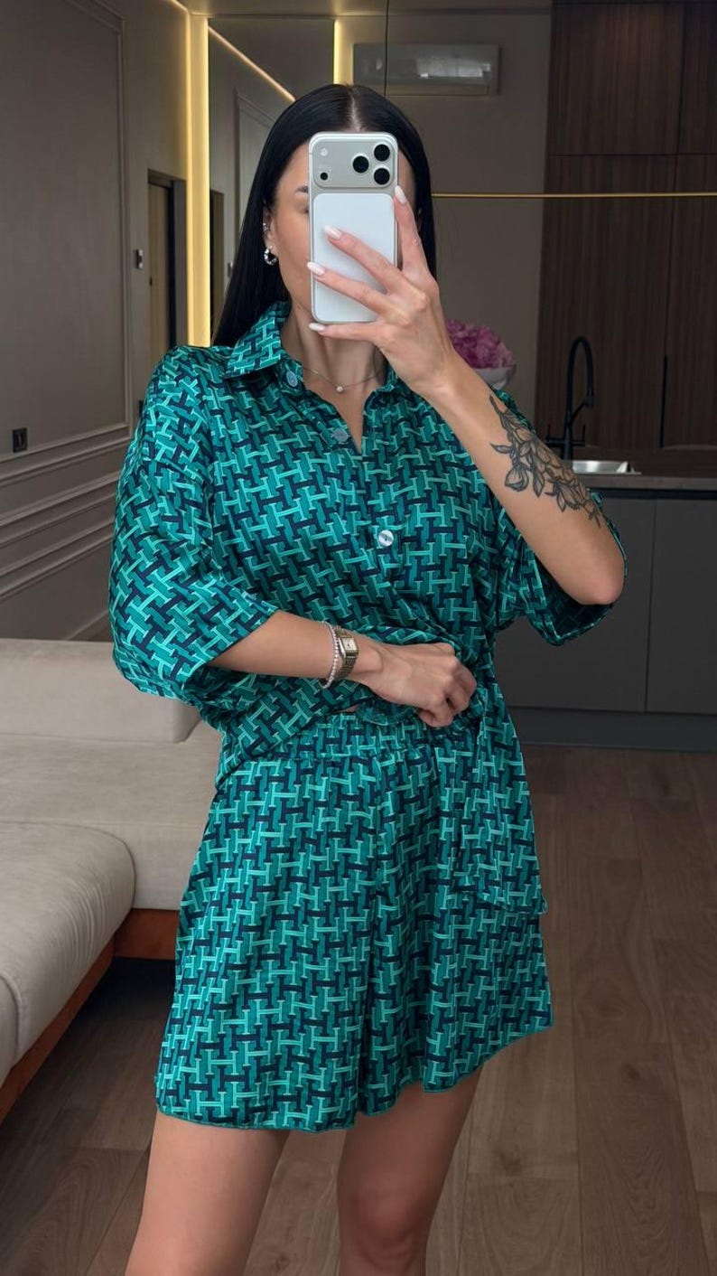 Puede incluir: Un conjunto de dos piezas compuesto por una camisa de manga corta con botones y pantalones cortos a juego. La tela presenta un patr&oacute;n geom&eacute;trico repetitivo en tonos verde oscuro, azul verdoso y negro, con un cuello cl&aacute;sico.