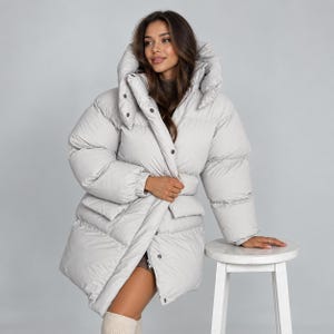 May include: A light gray puffer jacket featuring a high collar, a hood, long sleeves with elastic cuffs, and a front closure with visible snap buttons. The jacket has a quilted design with horizontal baffles and two front pockets at the waist.