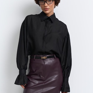 May include: A black shirt with flared sleeves and a burgundy leather mini skirt, worn with a black belt and tights. The shirt has a classic collar and the skirt is high-waisted. The belt has a gold buckle.