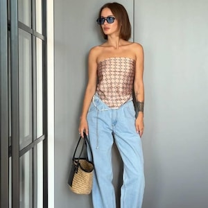Puede incluir: Un top beige sin tirantes con un patr&oacute;n geom&eacute;trico marr&oacute;n oscuro repetitivo, pantalones vaqueros azul claro, un bolso tipo cubo de paja con asas de cuero negro y sandalias de tac&oacute;n doradas con tiras. El conjunto incluye un cintur&oacute;n de cadena plateado.
