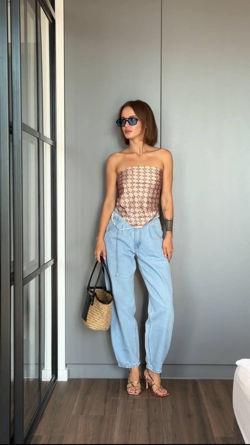 Puede incluir: Un top beige sin tirantes con un patr&oacute;n geom&eacute;trico marr&oacute;n oscuro repetitivo, pantalones vaqueros azul claro, un bolso tipo cubo de paja con asas de cuero negro y sandalias de tac&oacute;n doradas con tiras. El conjunto incluye un cintur&oacute;n de cadena plateado.