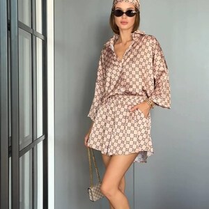 Puede incluir: Un conjunto de dos piezas en color beige y marr&oacute;n con estampado, compuesto por una camisa abotonada de manga tres cuartos y pantalones cortos a juego con bolsillos laterales. Se combina con un pa&ntilde;uelo, gafas de sol ovaladas y sandalias doradas.