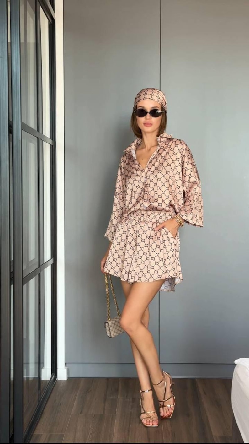 Puede incluir: Un conjunto de dos piezas en color beige y marr&oacute;n con estampado, compuesto por una camisa abotonada de manga tres cuartos y pantalones cortos a juego con bolsillos laterales. Se combina con un pa&ntilde;uelo, gafas de sol ovaladas y sandalias doradas.