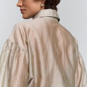 May include: A beige shirt with a collar and long sleeves, gathered at the cuffs. The fabric has a slight sheen. The model is wearing silver earrings. The background is plain white.
