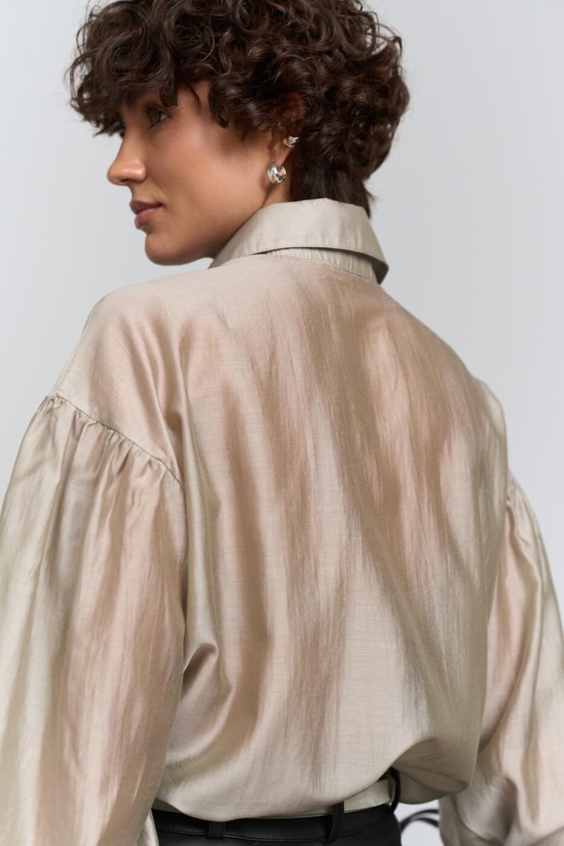 May include: A beige shirt with a collar and long sleeves, gathered at the cuffs. The fabric has a slight sheen. The model is wearing silver earrings. The background is plain white.