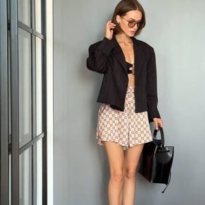 Puede incluir: Un blazer negro de rayas diplom&aacute;ticas sobre un bralette negro, combinado con pantalones cortos beige con un patr&oacute;n de monograma marr&oacute;n oscuro repetido. El conjunto incluye un bolso negro estructurado y sandalias de tiras doradas con tac&oacute;n.