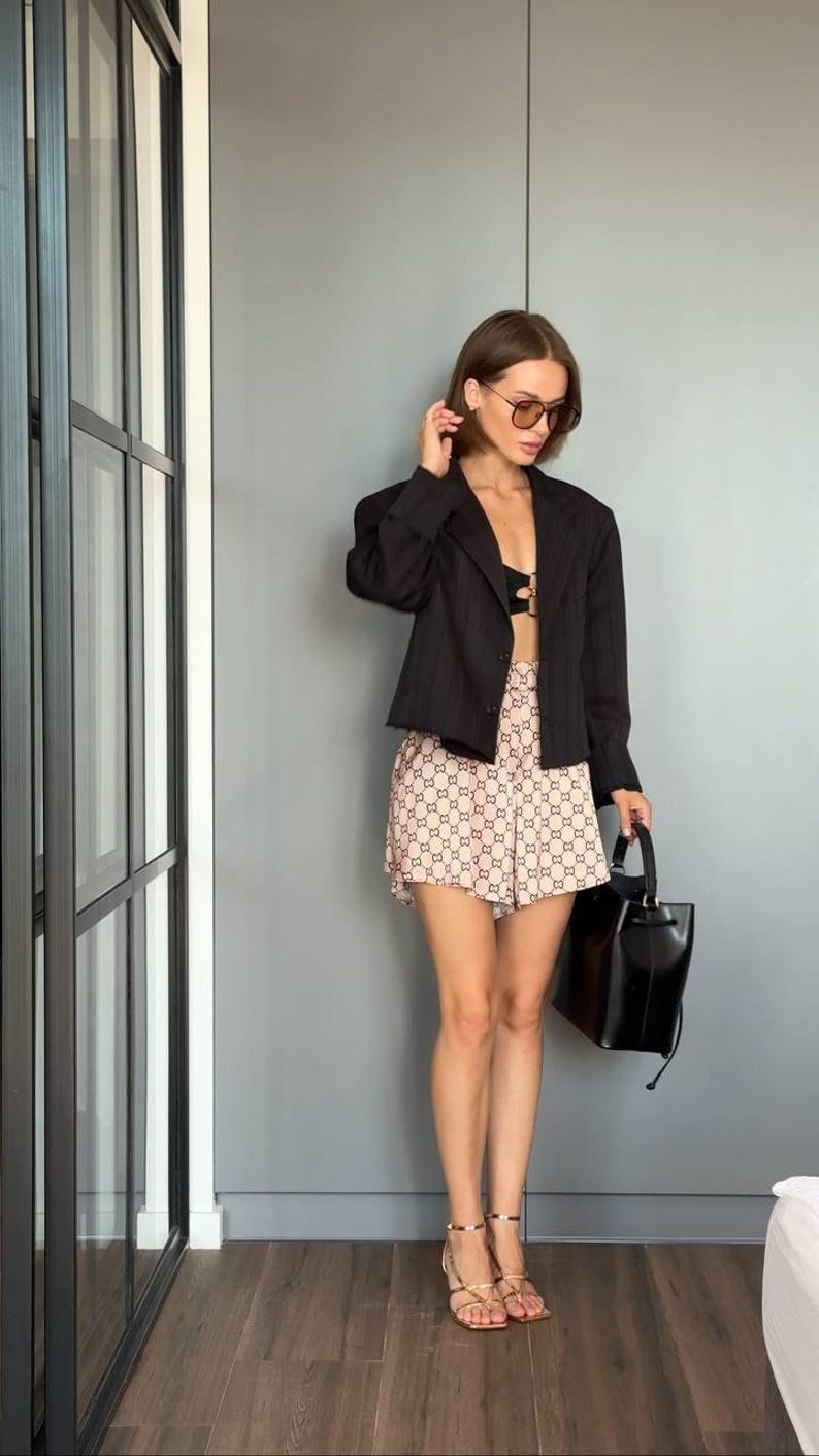 Puede incluir: Un blazer negro de rayas diplom&aacute;ticas sobre un bralette negro, combinado con pantalones cortos beige con un patr&oacute;n de monograma marr&oacute;n oscuro repetido. El conjunto incluye un bolso negro estructurado y sandalias de tiras doradas con tac&oacute;n.