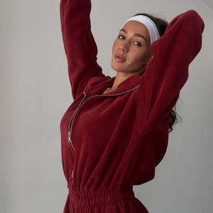 Może przedstawiać: Bordowy dres z polaru, składający się z bluzy z kapturem zapinanej na zamek i pasujących spodni ze sznurkiem. Bluza z kapturem ma srebrny zamek błyskawiczny, a spodnie mają elastyczny pas. Noszona jest biała opaska na głowę.