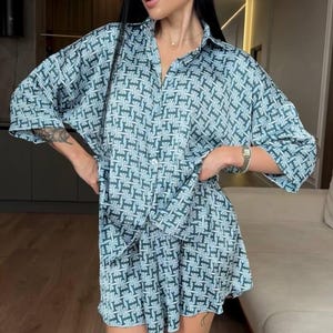 Puede incluir: Un conjunto de dos piezas compuesto por una camisa abotonada de manga al codo y pantalones cortos a juego. La prenda presenta un patr&oacute;n geom&eacute;trico repetitivo en tonos azul verdoso y azul claro, creando un look coherente con cuello cl&aacute;sico.