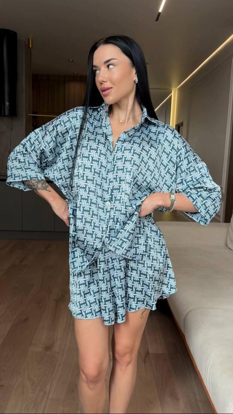 Puede incluir: Un conjunto de dos piezas compuesto por una camisa abotonada de manga al codo y pantalones cortos a juego. La prenda presenta un patr&oacute;n geom&eacute;trico repetitivo en tonos azul verdoso y azul claro, creando un look coherente con cuello cl&aacute;sico.