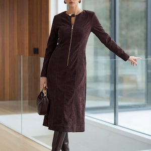 Pode incluir: Um vestido castanho escuro de manga comprida com fecho de correr comprido e bainha ao nível do joelho. O vestido é usado com botas altas a condizer e uma pequena mala. A modelo usa joias douradas.