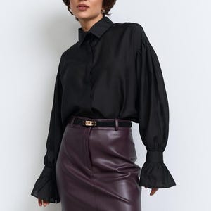 May include: A black shirt with long sleeves and frilled cuffs is worn with a burgundy leather mini skirt. A black belt with a gold buckle is at the waist. The outfit is finished with sheer black tights.