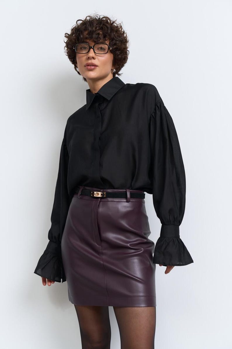 May include: A black shirt with long sleeves and frilled cuffs is worn with a burgundy leather mini skirt. A black belt with a gold buckle is at the waist. The outfit is finished with sheer black tights.