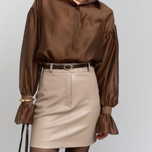May include: A brown blouse with flared sleeves, a beige leather skirt, and a brown belt. The outfit is accessorized with a gold bracelet and a brown handbag. The model is wearing black tights. The outfit is a stylish ensemble.