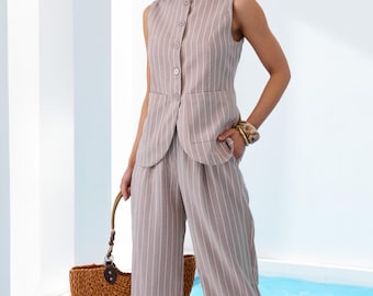 May include: A woman wearing a beige and white striped sleeveless vest, matching wide-leg pants, and a straw hat. She carries a woven handbag. The outfit is a neutral color, suitable for warm weather. The vest has button closures.