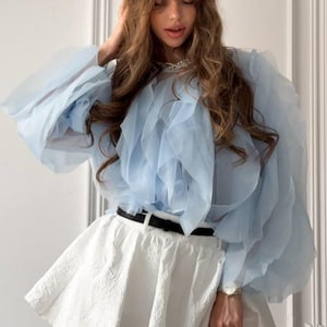 May include: A light blue, ruffled blouse with long, voluminous sleeves. The blouse is paired with a white, textured mini skirt and a black belt. A watch is visible on the wrist.