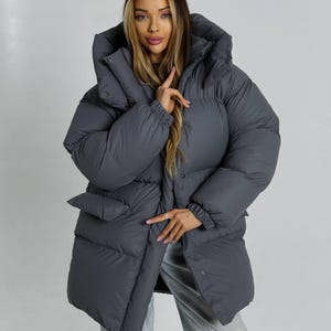May include: A charcoal gray oversized puffer jacket featuring a high collar, a large hood, long sleeves with elasticated cuffs, and two front flap pockets. The jacket is constructed with horizontal quilted panels and secures with a front snap button closure.