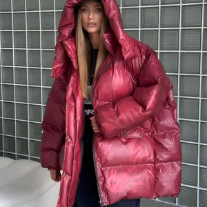 May include: A burgundy puffer jacket with a large hood. The jacket has a shiny, quilted texture and a zipper closure. The model is wearing a black cap and a black shirt. The background is a grid of square glass blocks.