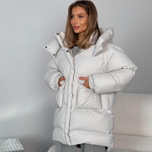 Puede incluir: Una chaqueta acolchada de color claro y de gran tamaño con una gran capucha. La chaqueta tiene una longitud larga, bolsillos y cierre de botón. El modelo lleva pantalones grises. La chaqueta es de color neutro, adecuada para varios conjuntos.