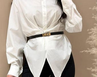 Puede incluir: Una camisa blanca abotonada con un diseño asimétrico único, combinada con pantalones negros y un cinturón negro con hebilla dorada. Un clutch negro se sostiene en la mano izquierda. El atuendo es elegante y moderno.