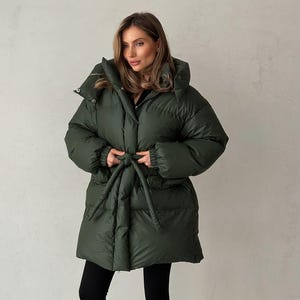 Può includere: Un cappotto imbottito verde oliva con cappuccio e cintura annodata. Il cappotto è lungo fino al ginocchio e ha un design trapuntato. La modella indossa leggings neri. Un capospalla elegante per il freddo.