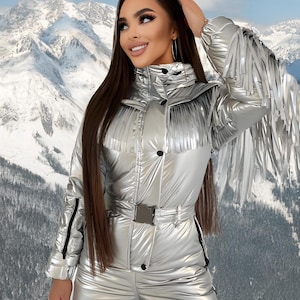 May include: Silver metallic snowsuit with a high collar, fringe detailing on the sleeves, and a belted waist. The one-piece suit has a shiny, quilted texture and black zippers. The background features a snowy mountain range.