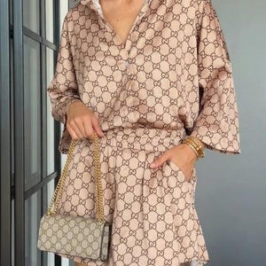 Puede incluir: Un conjunto de camisa abotonada y pantalones cortos a juego en color beige con un patr&oacute;n repetitivo de logotipos entrelazados en marr&oacute;n oscuro. Se combina con un peque&ntilde;o bolso de hombro rectangular con cadena dorada y una diadema a juego.