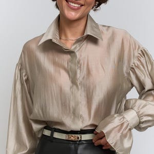 May include: A beige blouse with a collared neckline and long sleeves, paired with black leather trousers and a light-coloured belt with a gold buckle. The blouse has a slightly sheer appearance. The style is elegant and sophisticated.