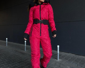 Puede incluir: Mono de nieve acolchado de color rosa brillante, con cinturón negro y ribetes negros. El mono de nieve tiene una capucha con ribete de piel sintética negra y cremallera frontal. La persona lleva gafas de sol negras y botas negras.