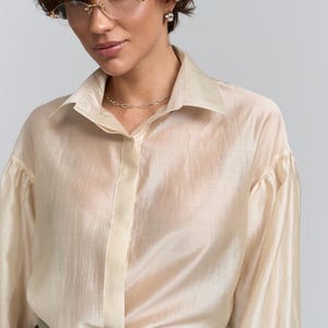 May include: A pale beige button-down shirt with long sleeves and a collared neckline. The shirt has a slightly sheer appearance. A pale green belt with a gold buckle is visible. The shirt is paired with black trousers.