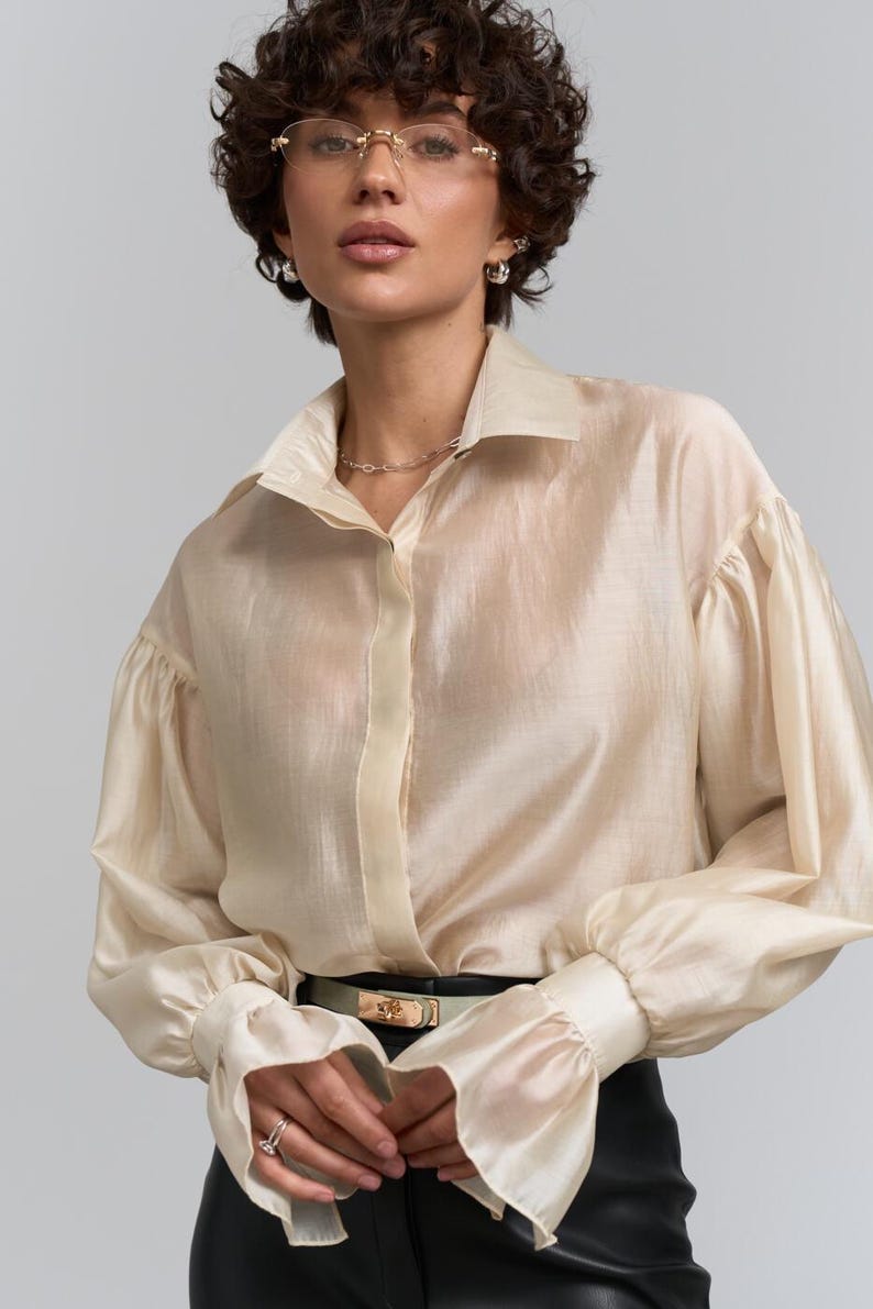 May include: A cream-coloured blouse with long, voluminous sleeves and a classic collar. The blouse is paired with a black belt and trousers. The fabric appears to be sheer and lightweight, creating an elegant look.