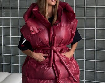 May include: A burgundy puffer vest with a large hood and tie waist. The vest has a quilted design and is worn over a black shirt and paired with navy blue pants. A black baseball cap completes the look.