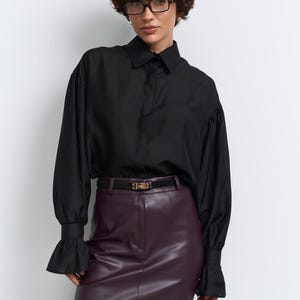 May include: A black shirt with flared sleeves and a burgundy leather mini skirt, worn with a black belt and tights. The shirt has a classic collar and the skirt is high-waisted. The belt has a gold buckle.