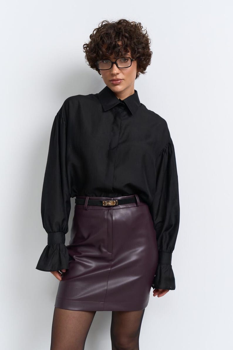 May include: A black shirt with flared sleeves and a burgundy leather mini skirt, worn with a black belt and tights. The shirt has a classic collar and the skirt is high-waisted. The belt has a gold buckle.