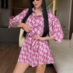 Puede incluir: Un vestido camisero con estampado rosa y blanco y un pa&ntilde;uelo a juego. El vestido presenta cuello camisero, mangas hasta el codo y un cintur&oacute;n para anudar a la cintura. La tela muestra un motivo geom&eacute;trico repetitivo de la letra H en toda la prenda.