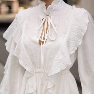 K&ouml;nnte beinhalten: Ein wei&szlig;es Kleid mit hohem R&uuml;schenkragen und langen &Auml;rmeln. Das Kleid hat einen Schl&uuml;sselloch-Ausschnitt mit Krawattenverschluss und gestufte R&uuml;schen, die &uuml;ber das Oberteil fallen. Das Kleid ist aus einem strukturierten Stoff gefertigt.